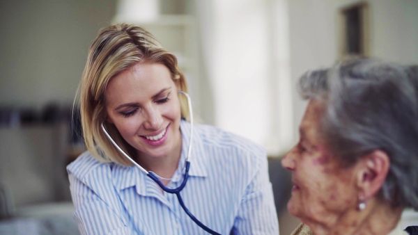 A young health visitor examining a senior woman with a stethoscope at home. Slow motion.