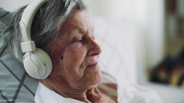 A close-up of sick senior woman with headphones lying in bed at home or in hospital, listening to music. Slow motion.