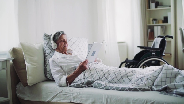 A close-up of sick senior woman with headphones and tablet lying in bed at home or in hospital, listening to music. Slow motion.