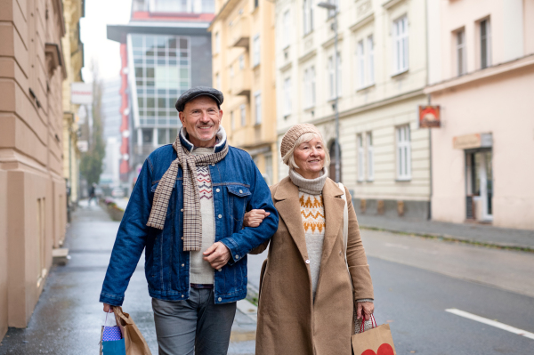 Portrait of happy senior couple walking outdoors on street in city, carrying shopping bags.