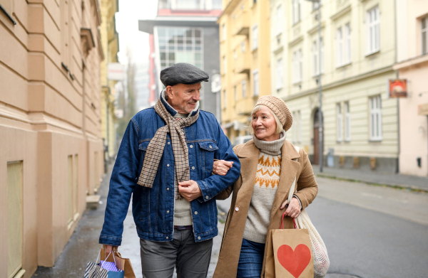 Portrait of happy senior couple walking outdoors on street in city, carrying shopping bags.