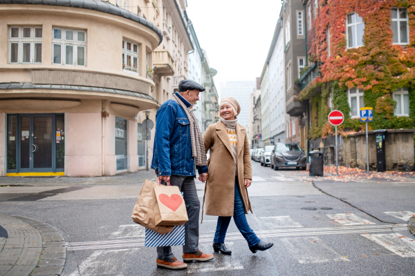 Portrait of happy senior couple walking outdoors on street in city, carrying shopping bags.