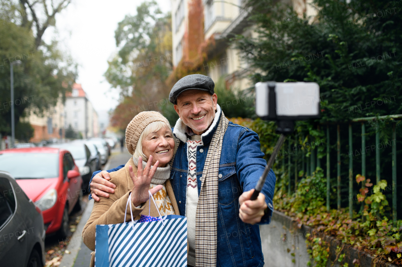 Portrait of happy senior couple walking outdoors on street in city, taking selfie.