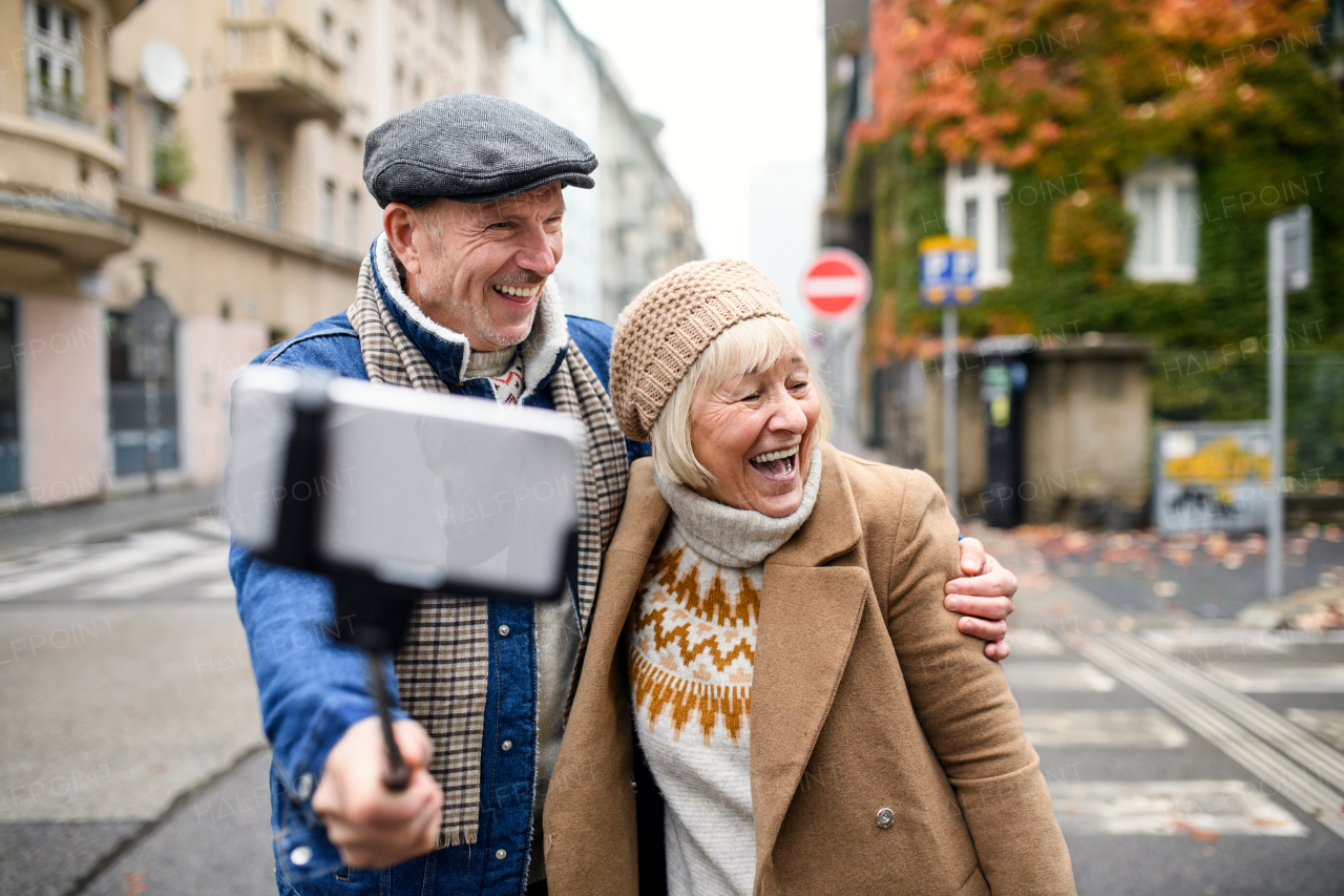 Portrait of happy senior couple walking outdoors on street in city, taking selfie.