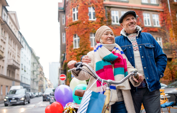 Portrait of happy senior couple with bicycle and balloons outdoors on street in city.