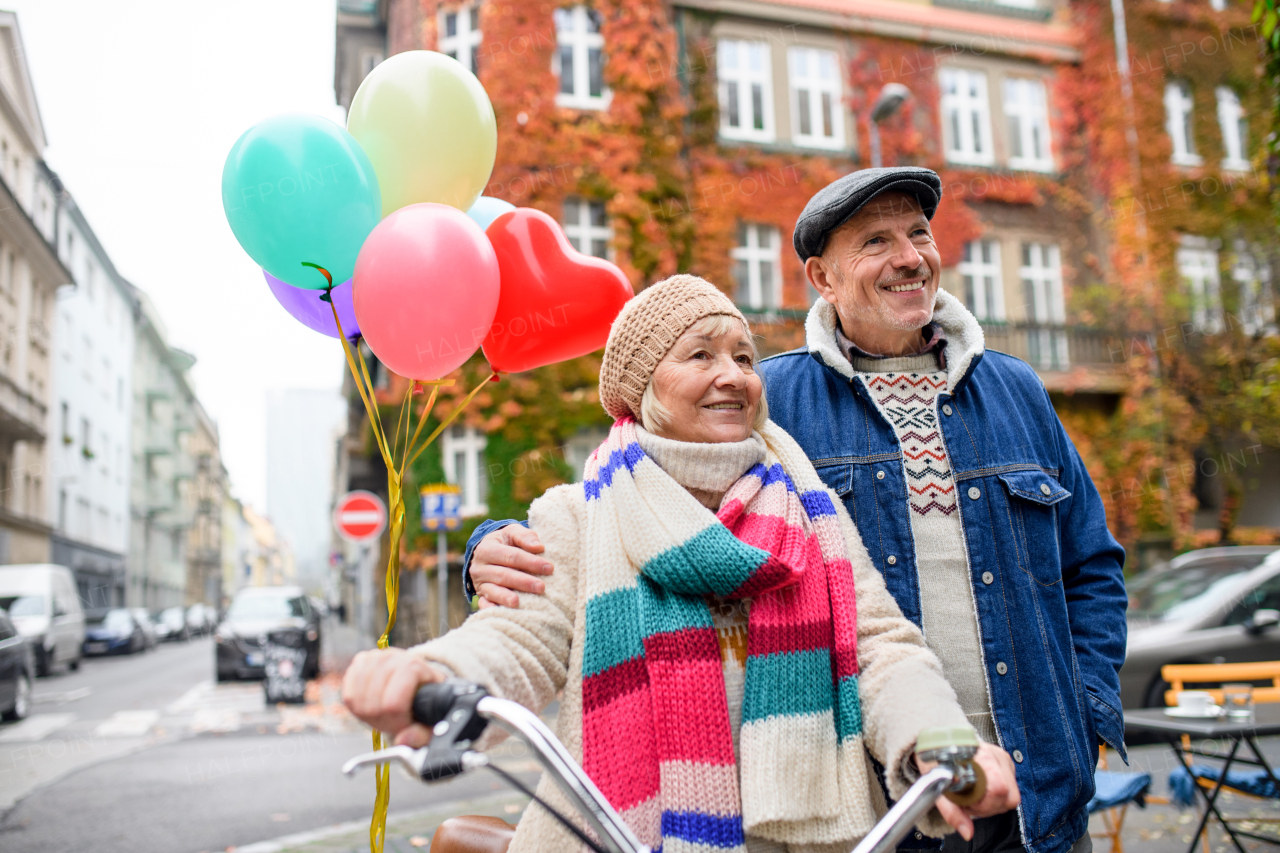 Portrait of happy senior couple with bicycle and balloons outdoors on street in city.