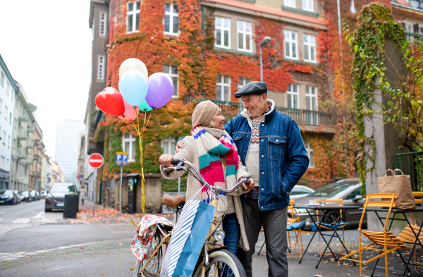 Portrait of happy senior couple with bicycle and balloons outdoors on street in city.