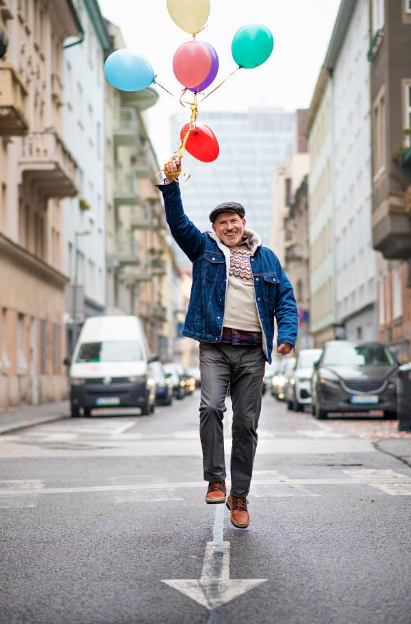 Happy senior man with balloons outdoors on street in city, walking. Party concept.