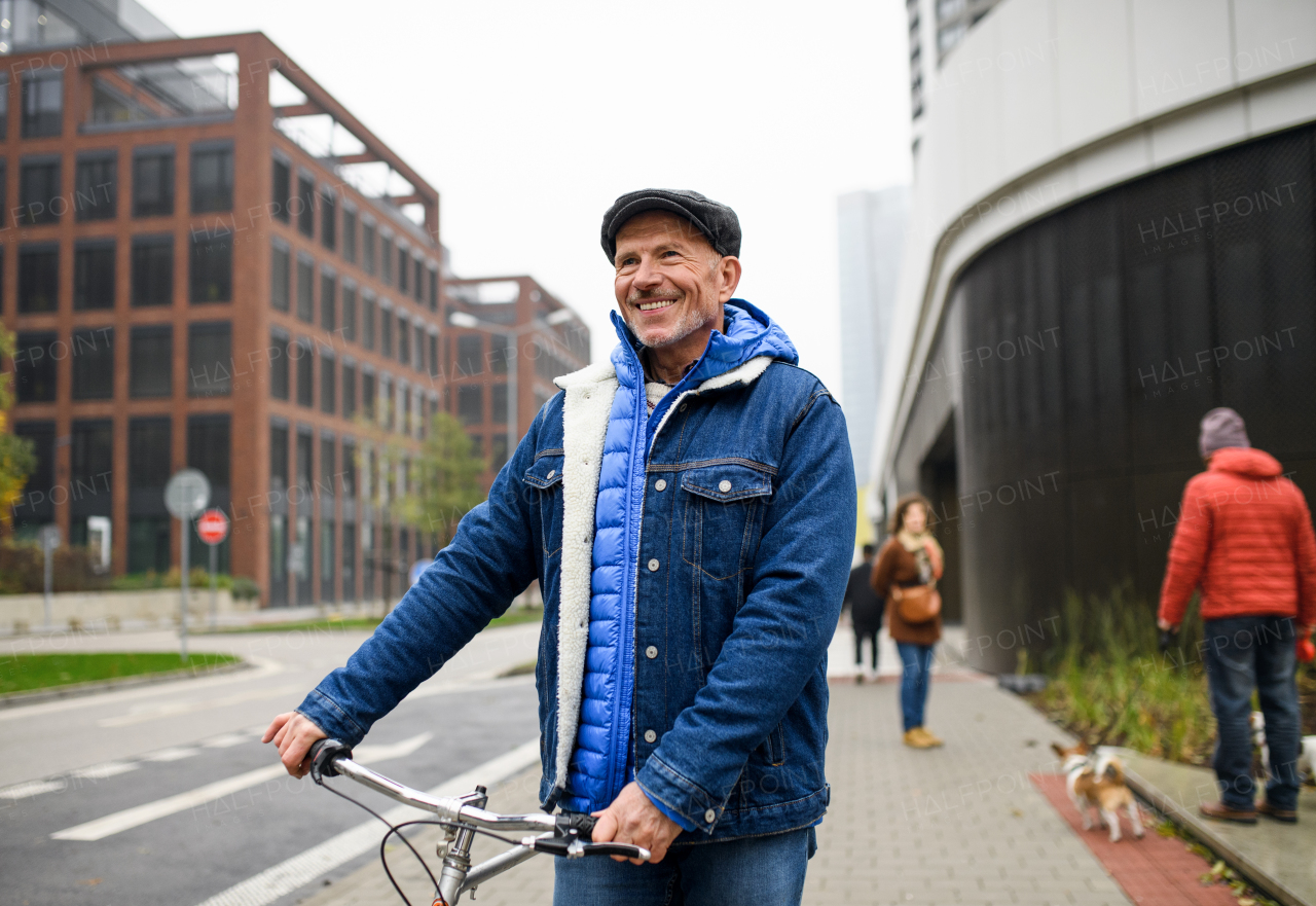 Happy senior man commuter with bicycle outdoors on street in city, walking.