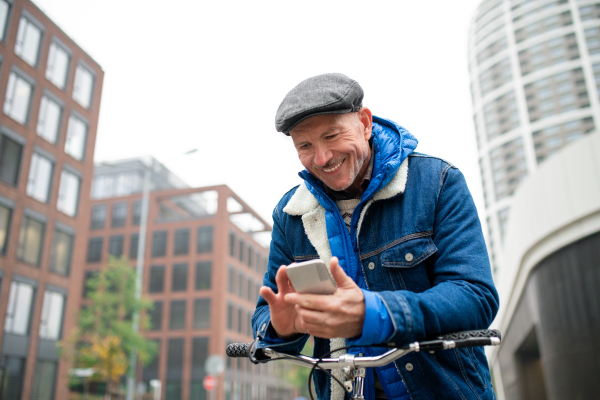 Happy senior man commuter with bicycle outdoors on street in city, using smartphone.