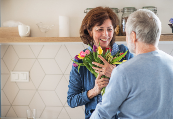 A senior couple in love indoors at home, man giving flowers to happy woman.