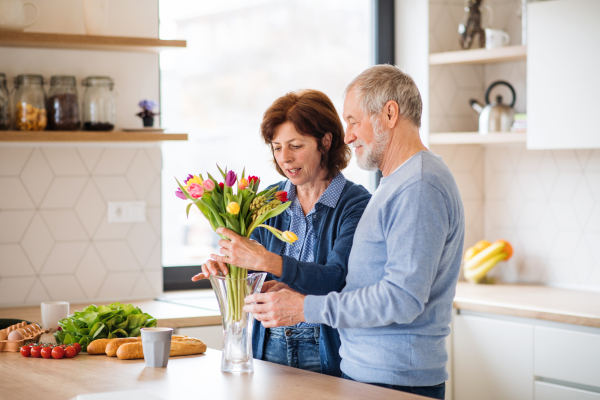 A portrait of senior couple in love putting flowers in vase indoors at home, unpacking shopping.