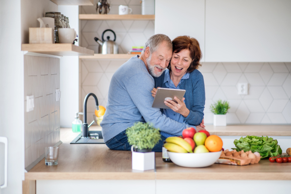 A portrait of happy senior couple in love indoors at home, looking for recipe using tablet.