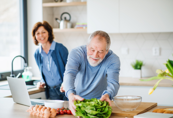 A portrait of happy senior couple with laptop indoors at home, cooking.