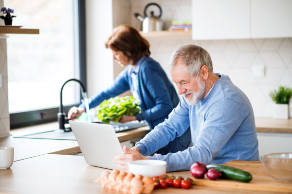 A portrait of happy senior couple with laptop indoors at home, cooking.