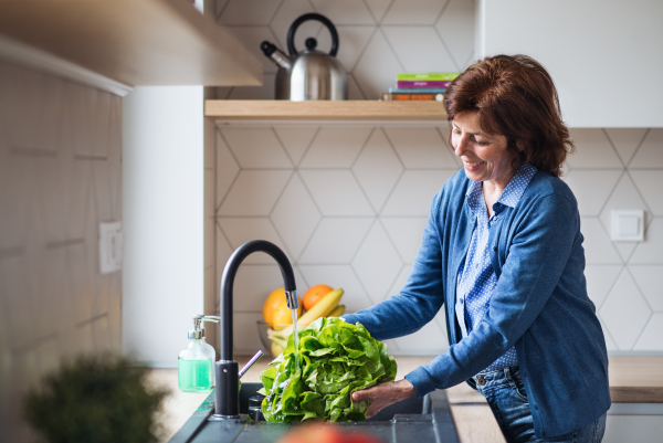 A portrait of happy senior woman indoors at home, washing lettuce vegetable.