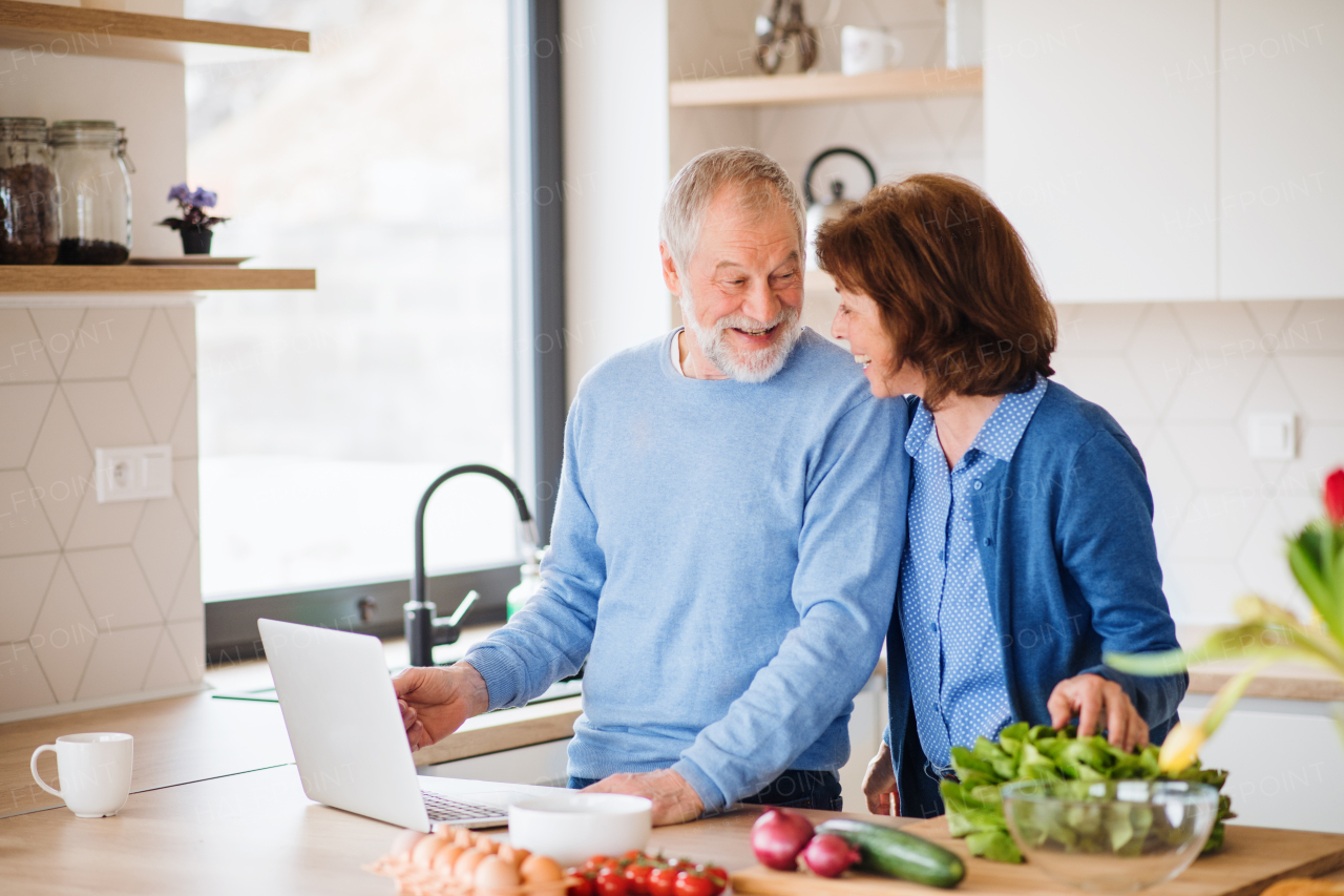 A portrait of happy senior couple with laptop indoors at home, cooking.