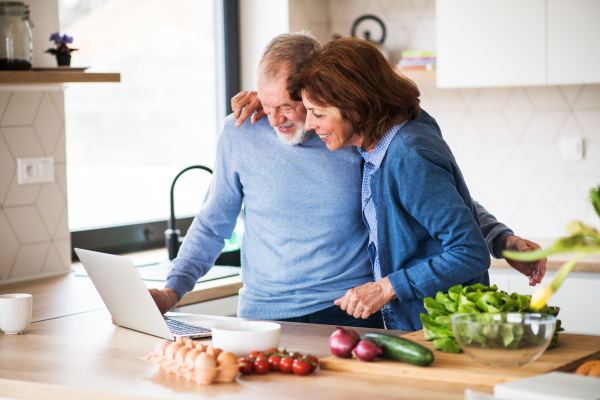 A portrait of happy senior couple with laptop indoors at home, cooking.