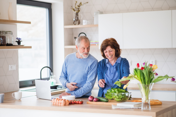 A portrait of happy senior couple with laptop indoors at home, cooking.