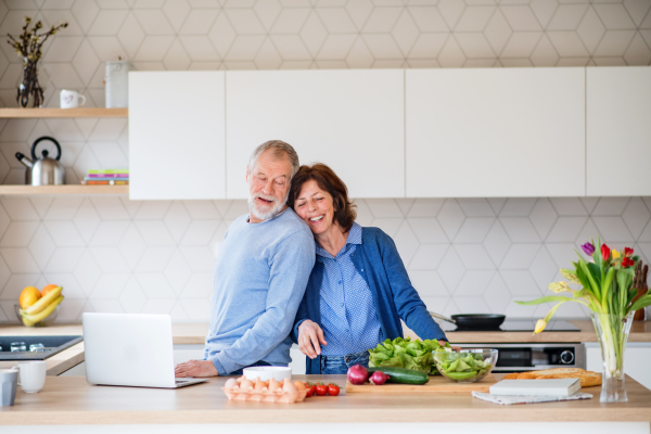 A portrait of happy senior couple with laptop indoors at home, cooking.