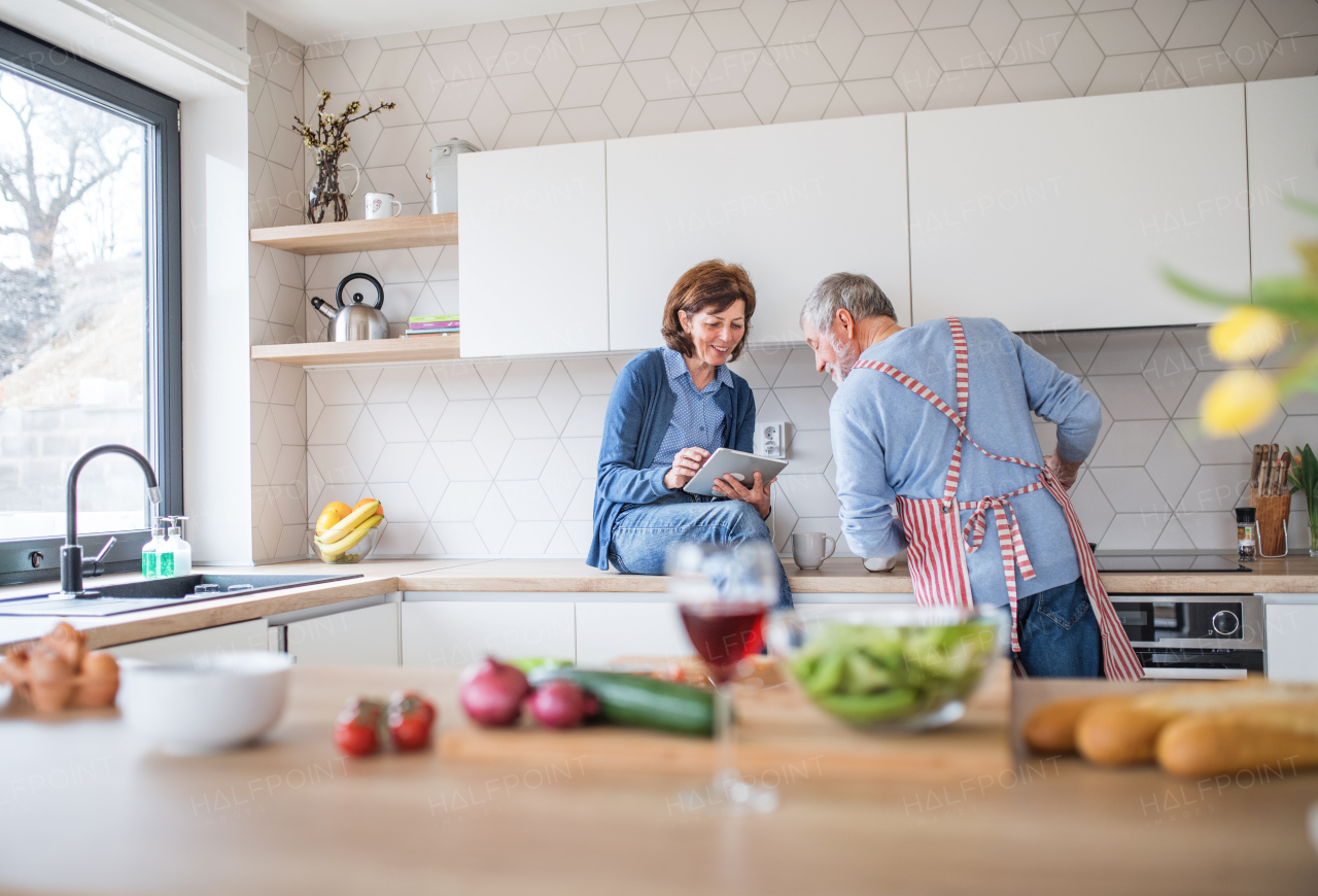 A portrait of happy senior couple in love indoors at home, looking for recipe using tablet.