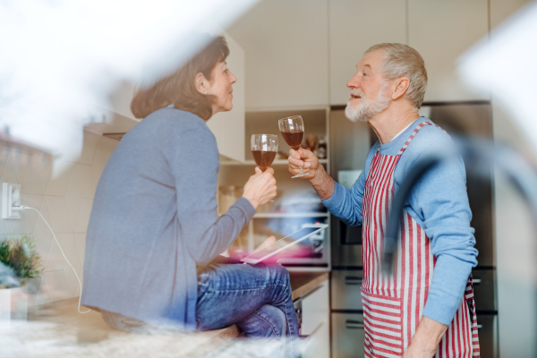 A portrait of happy senior couple in love indoors at home, clinking glasses. Shot through glass.