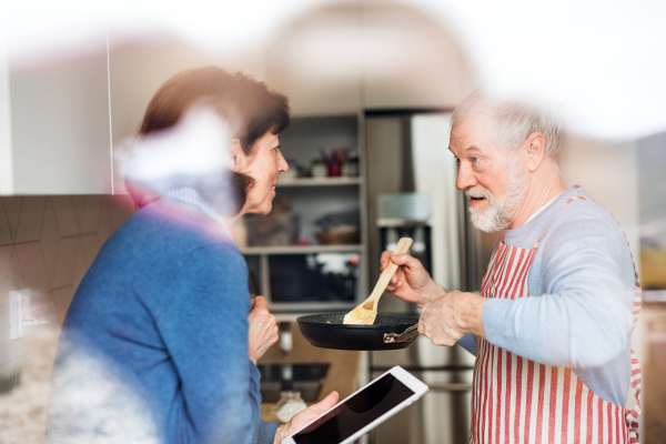 A portrait of happy senior couple in love indoors at home, cooking. Shot through glass.