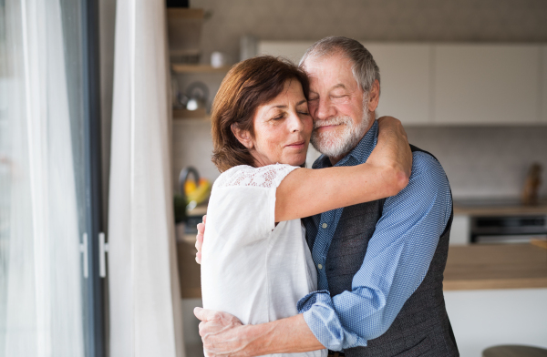 An affectionate senior couple in love standing indoors at home, hugging.