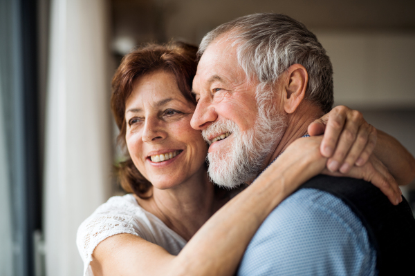 A senior couple standing indoors at home, looking out of window.