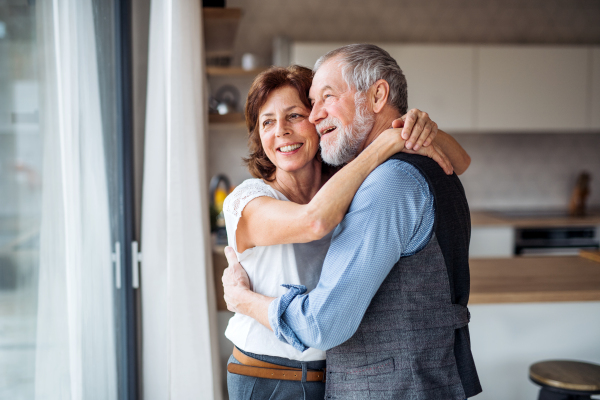 A senior couple standing indoors at home, looking out of window.