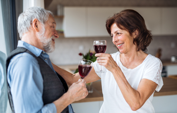 A portrait of happy senior couple in love indoors at home, clinking glasses.