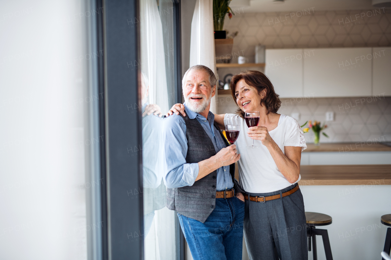 A senior couple with wine standing indoors at home, looking out of window.