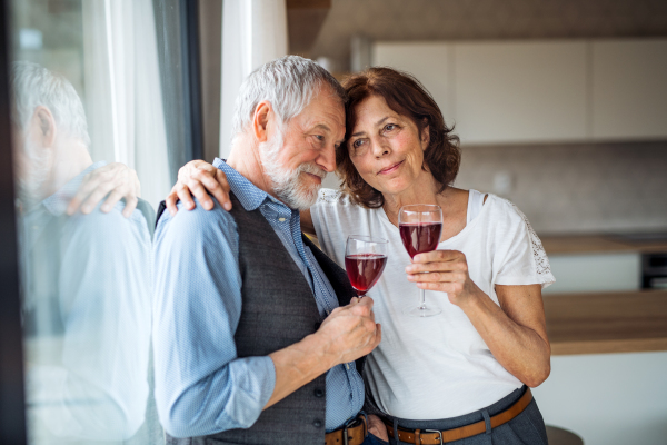 A senior couple with wine standing indoors at home, standing by window.