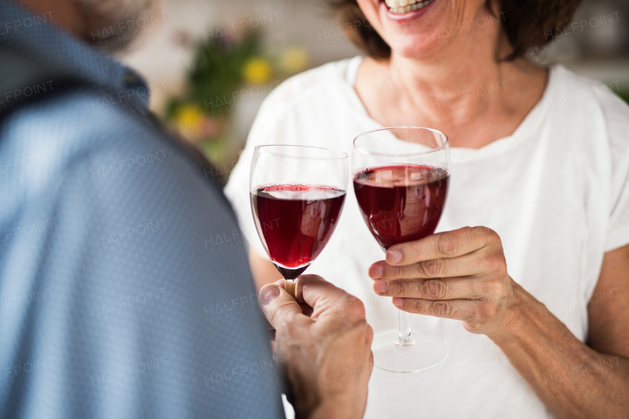 A midsection of senior couple in love indoors at home, clinking glasses with red wine.