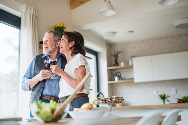 A senior couple with wine standing indoors at home, looking out of window.