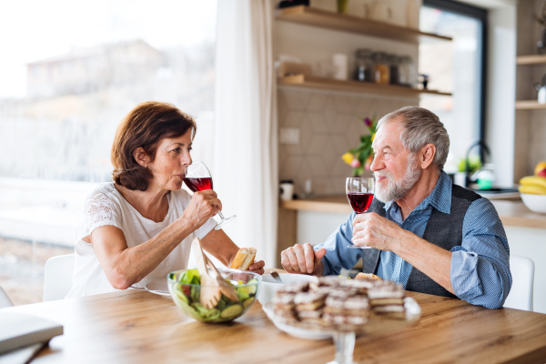 A senior couple with wine having lunch indoors at home, talking.