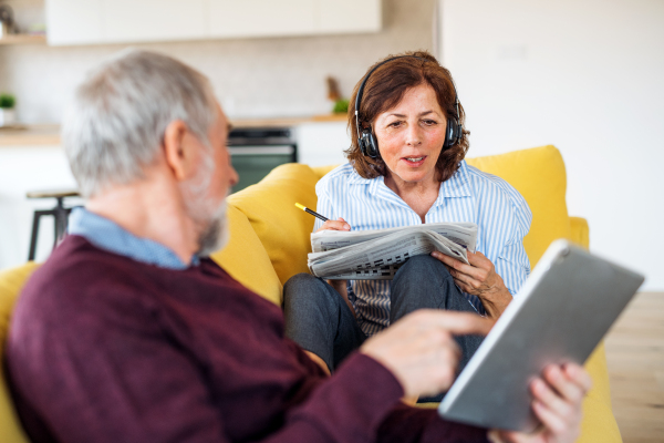 Senior couple with tablet and headphones sitting on sofa indoors at home, relaxing.