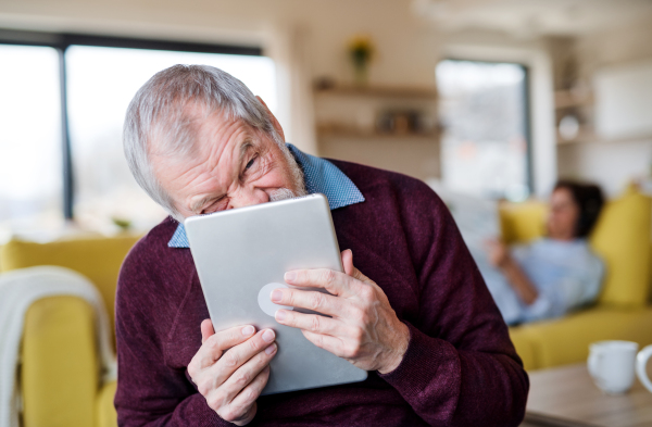 Front view of senior man with tablet indoors at home, making funny faces.