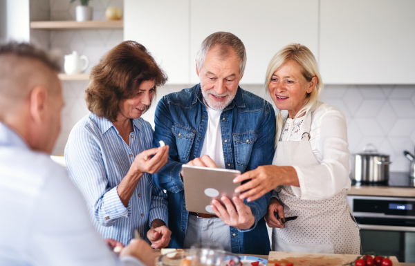 Front view of group of cheerful senior friends with tablet at dinner party at home, cooking.