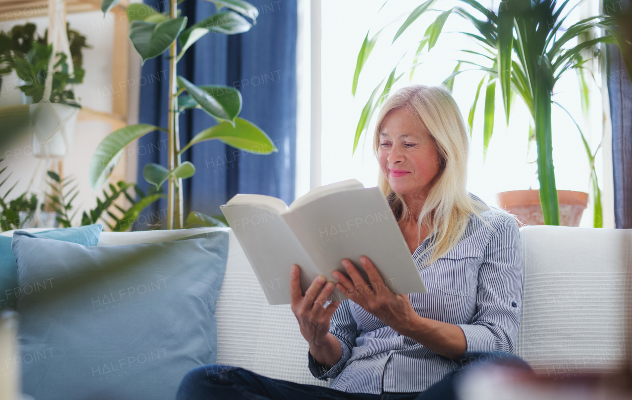 Front view of ttractive senior woman sitting indoors on sofa, reading book and relaxing.