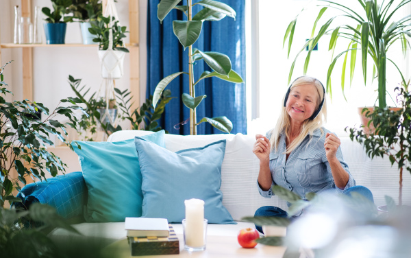 Happy attractive senior woman with headphones sitting indoors on sofa, relaxing.