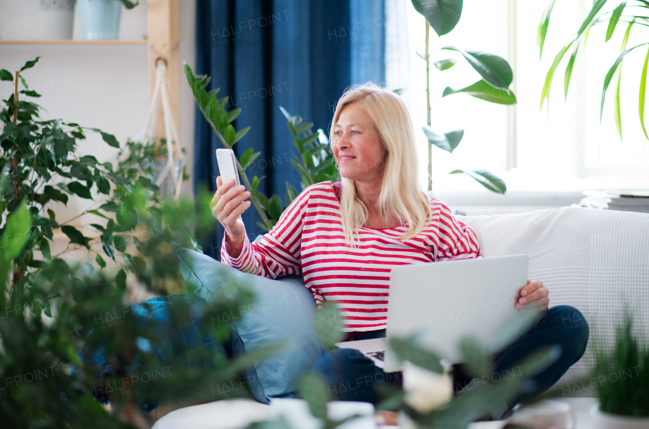 Attractive senior woman with laptop and smartphone sitting indoors on sofa, taking selfie.