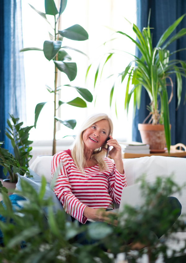 Happy attractive senior woman with smartphone sitting indoors on sofa, making phone call.