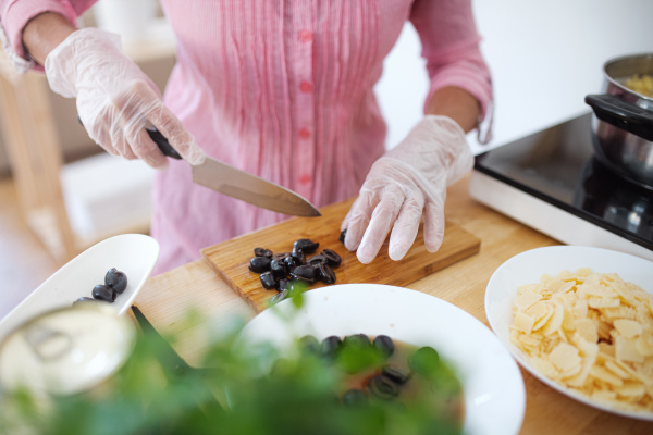 Unrecognizable woman with gloves cooking in kitchen indoors, chopping.