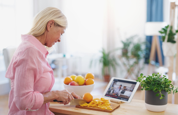 Attractive senior woman preparing food in kitchen indoors, following food vlogger on tablet.