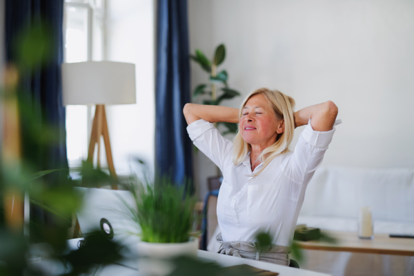 Attractive senior businesswoman with laptop sitting indoors at desk in home office, stretching when working.