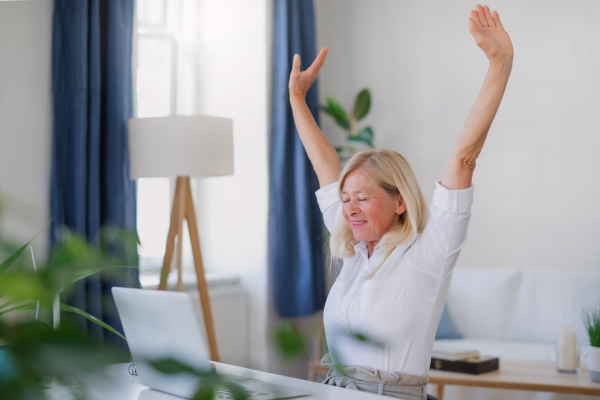 Attractive senior businesswoman with laptop sitting indoors at desk in home office, stretching when working.