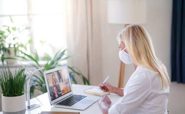 Side view of senior businesswoman with face mask indoors in home office, having video call.