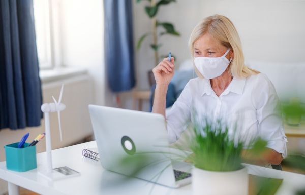 Senior woman engineer with face mask and laptop sitting indoors in office, working.