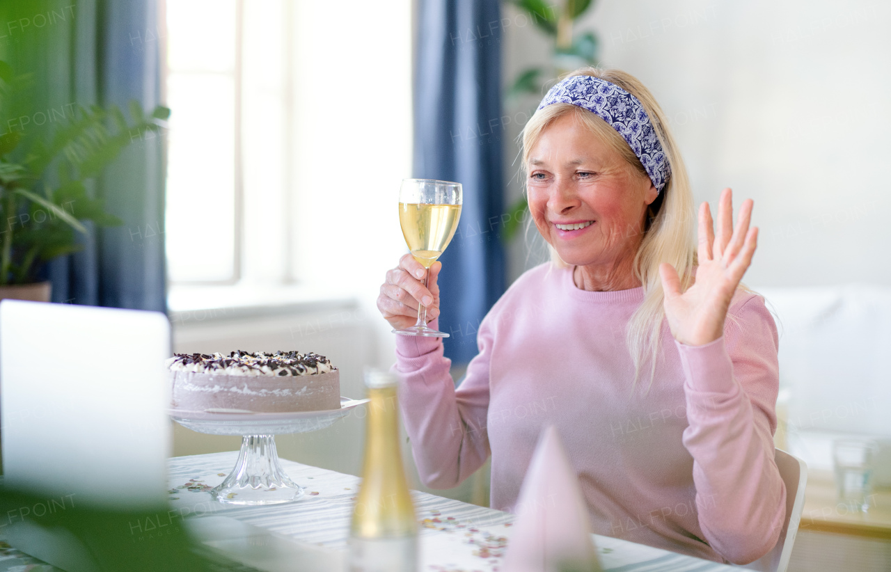 Senior woman with laptop, cake and wine indoors at home, celebrating distance birthday.